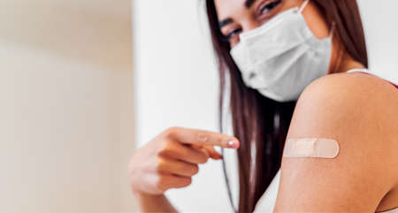 Young girl woman showing pointing with finger to bandage on her arm shoulder wearing protective mask done with vaccination. Female just got vaccinated against Covid19 face mask looking at camera.の写真素材