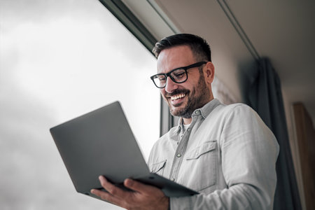 Portrait of young adult smiling cheerful businessman entrepreneur in office using laptop computer having online video call conference meeting standing next to window.の写真素材