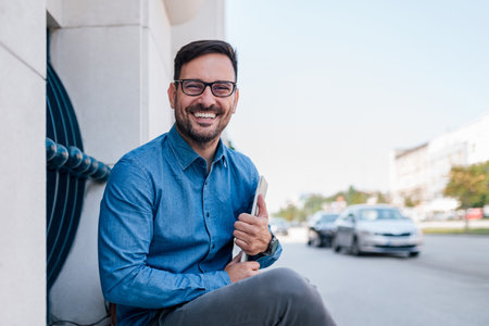 Portrait of smiling young businessman holding laptop. Cheerful male professional commuter in formals is sitting against building. He is commuting in the city.の写真素材