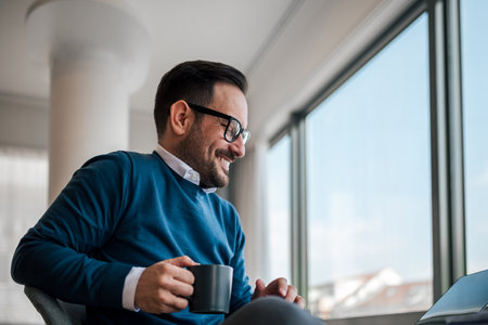 Smiling businessman having coffee while working. Young male professional is sitting at desk. He is wearing smart casuals in office.の写真素材