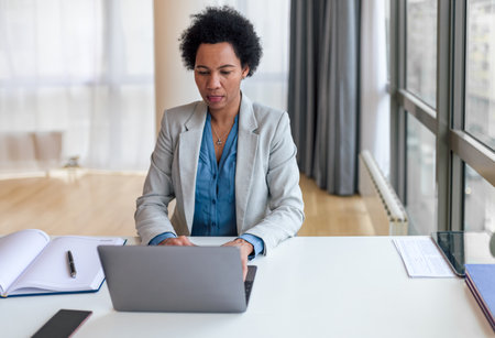 Confident businesswoman using laptop at desk. Female entrepreneur is sitting by window. She is wearing formals while working at office.の写真素材