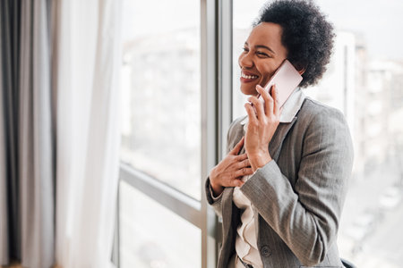 Smiling businesswoman talking on smart phone. Female executive is wearing formals. She is standing against window at office.の写真素材