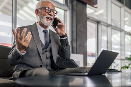 Happy manager gesturing and talking on cellphone while using laptop at office.の写真素材
