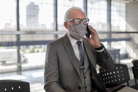 Senior businessman wearing mask talking on smart phone. Elderly male entrepreneur is waiting at subway station during corona virus pandemic. He is following protective measures.の写真素材