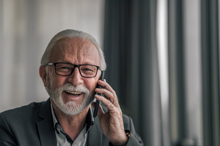 Portrait of business professional talking on mobile phone. Senior male entrepreneur is working on laptop at desk. He wearing formals at corporate workplace.の写真素材