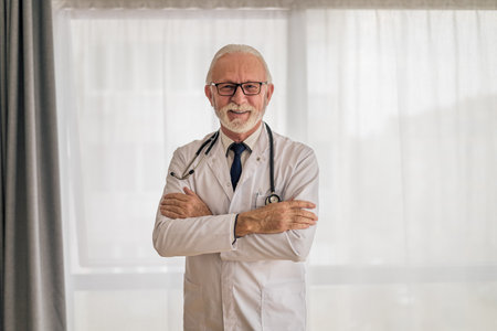Portrait of confident smiling elderly healthcare professional, Senior male doctor is standing with arms crossed against window, He is wearing lab coat and stethoscope.の写真素材