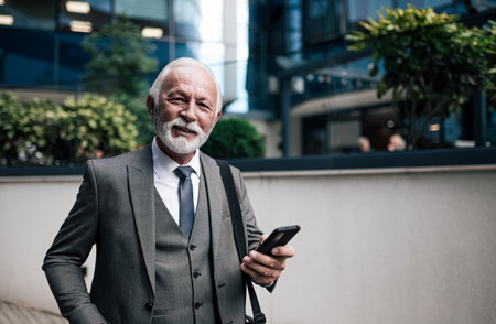 Portrait of smiling elderly businessman using smart phone. Elderly male professional wearing suit is carrying laptop bag in the city. He is standing against modern office building during travel.の写真素材