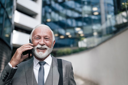 Close-up of smiling senior businessman discussing on smart phone. Elderly male professional with bag standing against office building. He is wearing suit while traveling in the city.の写真素材