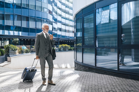 Elderly male professional with suitcase talking through speaker on mobile phone. Senior businessman wearing suit walking on footpath against office building in the city. He is carrying laptop bag.の写真素材