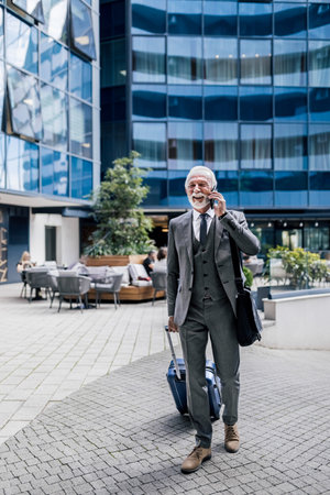 Smiling senior businessman with suitcase talking on mobile phone. Male professional wearing suit is carrying laptop bag. He is walking on footpath against office building while traveling in the city.の写真素材