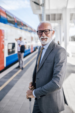 Portrait of smiling senior businessman wearing suit standing by train. Male entrepreneur with luggage is waiting at subway station platform. He is commuting during sunny day.の写真素材