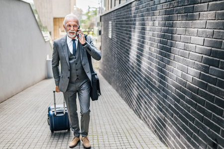 Senior businessman talking on smart phone. Elderly male professional wearing formals is carrying laptop bag. He is walking on footpath against office building in the city.の写真素材