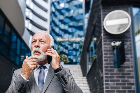 Confused senior entrepreneur or businessman talking on smart phone. Elderly professional is discussing while standing against office building in the city. He is wearing full suit.の写真素材