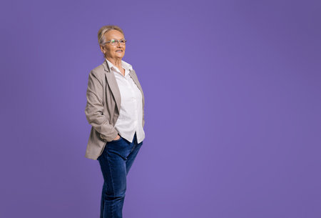 Confident senior female professional manager with hands in pockets looking away and contemplating while standing isolated over purple backgroundの写真素材
