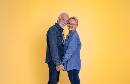 Side view of romantic senior couple with head to head holding hands affectionately and looking at camera isolated on yellow backgroundの写真素材