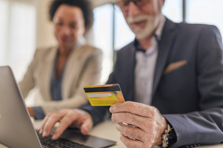 Close-up of senior businessman paying online through laptop. Elderly male executive is sitting with female coworker. Focus is on credit card at desk in office.の写真素材