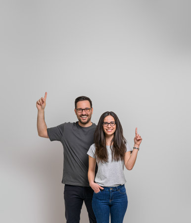 Smiling young couple pointing up at copy space for marketing against background. Happy man and woman wearing eyeglasses showing empty space for advertisementの写真素材