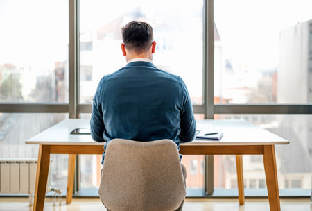 Rear view of businessman working at desk. Male professional is sitting in front of windows. He is wearing sweater at corporate office.の写真素材
