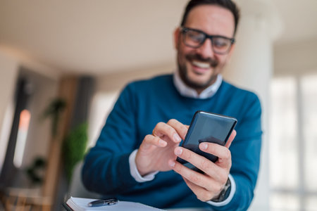 Close-up of young businessman text messaging on mobile phone. Smiling male executive is wearing smart casuals at office. Focus is on smart phone and hands..の写真素材