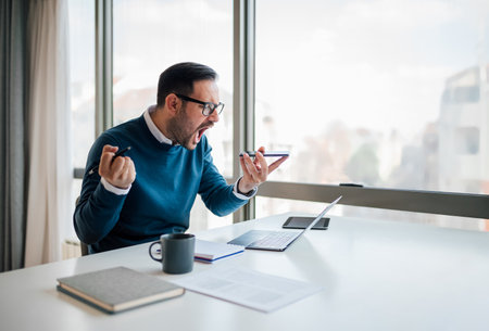 Angry young professional shouting while discussing on smart phone. Displeased business executive is working at desk by window. He is sitting in corporate office.の写真素材