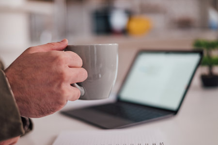 Cropped image of male executive holding coffee cup. Businessman working at desk in office. Blurred image of laptop in background.の写真素材