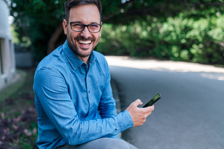 Portrait of smiling businessman using smart phone. Young male professional is wearing formals. He is sitting by road against trees at the city.の写真素材
