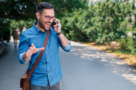 Charming adult man in blue shirt, having a conversation over his phone, talking to his friend while walking.の写真素材
