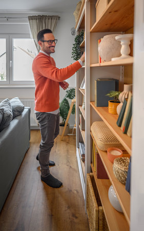 Side view of smiling male entrepreneur dressed in casuals searching book on shelf in home library. Young businessman choosing book for reading on bookshelf while working from home officeの写真素材