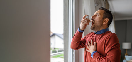 Sick young male patient touching chest in pain and inhaling through asthma inhaler by window at home