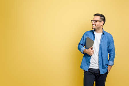Happy male freelancer with hand in pocket holding digital tablet looking away over yellow backgroundの写真素材