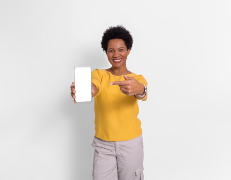 Smiling young woman pointing at blank phone screen and recommending apps on white backgroundの写真素材
