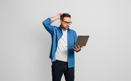 Confused disappointed businessman with hand in hair reading e-mail over laptop on gray backgroundの写真素材