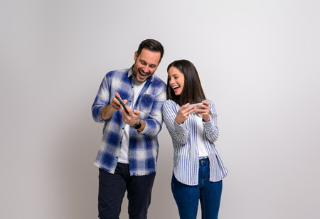 Cheerful young girlfriend playing video game and looking at boyfriend's mobile phone while competing him. Couple enjoying gaming together with cellphones while standing on backgroundの写真素材