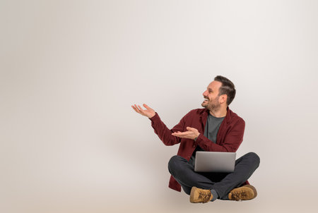 Happy male executive showing copy space for business marketing and sitting with legs crossed. Young salesman advertising something with hands while working online on laptop against backgroundの写真素材