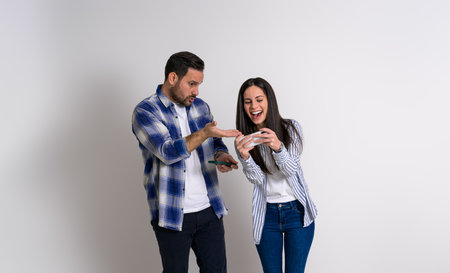 Displeased young man pointing at cheerful girlfriend's smart phone while playing video game against background. Couple dressed in casuals with cellphones gaming and competing each otherの写真素材