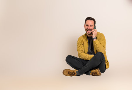 Young businessman dressed in casuals laughing and having business conversation over smart phone. Male professional cheerfully talking on cellphone while sitting with crossed legs against backgroundの写真素材