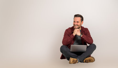 Smiling handsome male entrepreneur with hands clasped looking away while sitting with crossed legs. Young sales executive working over wireless computer and posing cheerfully on backgroundの写真素材