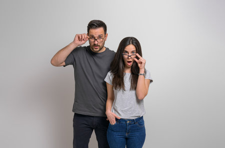 Amazed young couple with wide open eyes and mouth looking out eyeglasses at camera. Shocked man and woman dressed in casuals listening to unexpected news against backgroundの写真素材