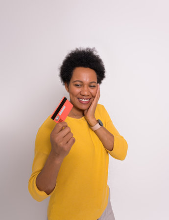 Smiling young woman touching chin and thinking while looking at credit card on white background