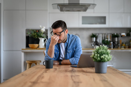 Exhausted and anxious businessman with head in hand sitting by laptop at desk in home officeの写真素材