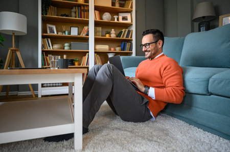Young smiling businessman doing research over laptop while relaxing by sofa on carpet in living roomの写真素材