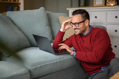 Smiling young businessman wearing eyeglasses checking e-mails over laptop on sofa in living roomの写真素材