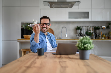 Cheerful male freelancer talking over speaker on smart phone while laptop on desk in home officeの写真素材