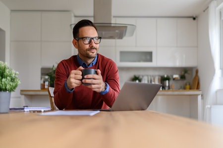 Young businessman holding coffee cup and contemplating while sitting with laptop on desk at homeの写真素材