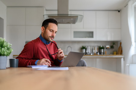Handsome entrepreneur holding glasses and thinking ideas while using laptop on desk in home officeの写真素材
