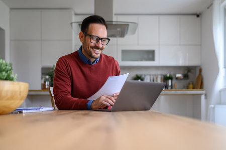 Smiling male financial adviser holding spreadsheets and analyzing report over laptop at home officeの写真素材
