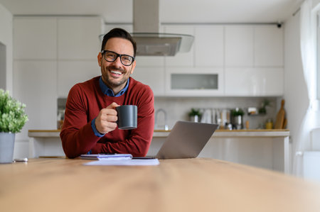 Portrait of smiling businessman holding coffee cup while sitting by laptop at desk in home officeの写真素材