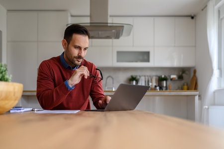Focused young businessman holding glasses and working online over laptop on desk in home officeの写真素材