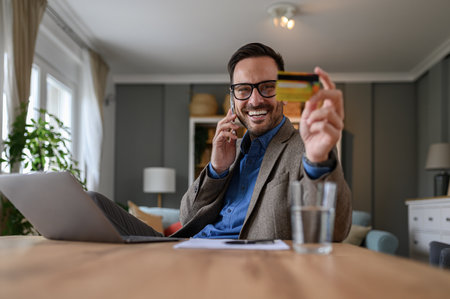 Handsome entrepreneur in elegant suit holding credit card and talking over smart phone at deskの写真素材