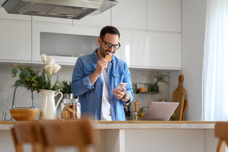 Happy young businessman eating cookie while checking messages over mobile phone at kitchen counterの写真素材